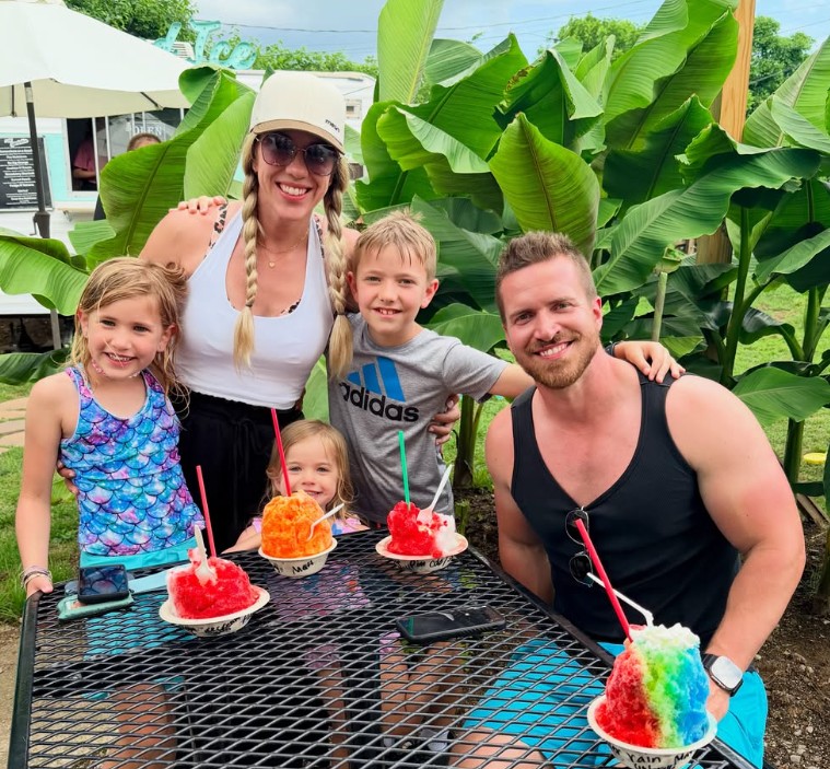 A family of five with rainbow shave ice at a shaded outdoor table framed by banana leaves