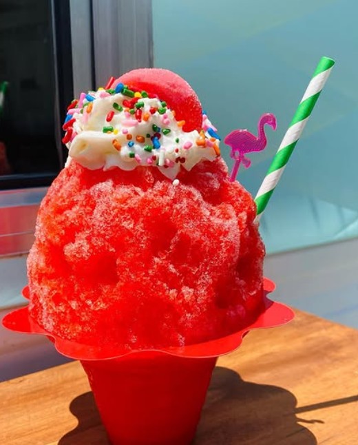 A strawberry shave ice topped with whipped cream, rainbow sprinkles and a flamingo pick